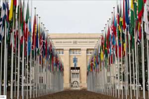The UN Palais des Nations in Geneva, a cream coloured stone building with an avenue of flags leading up to it.
