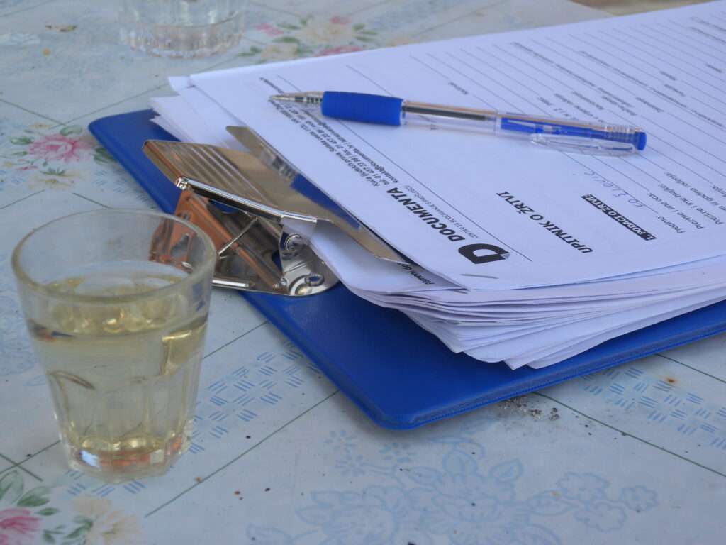 A close up of a form on a clipboard, sitting on a table with a pen on top and a glass to one side.