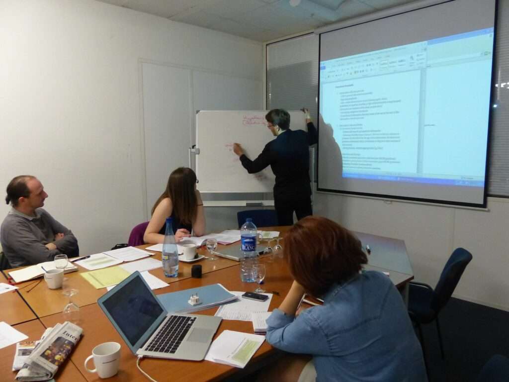 Two women and a man sit around a table covered with papers, laptops and cups. They watch another man who is writing on a whiteboard. To his right is a projector showing a Word document.