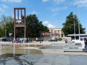 The UN Palais des Nations in Geneva, a cream coloured stone building with an avenue of flags leading up to it. Opposite this is a sculpture of a giant chair, and some fountains.